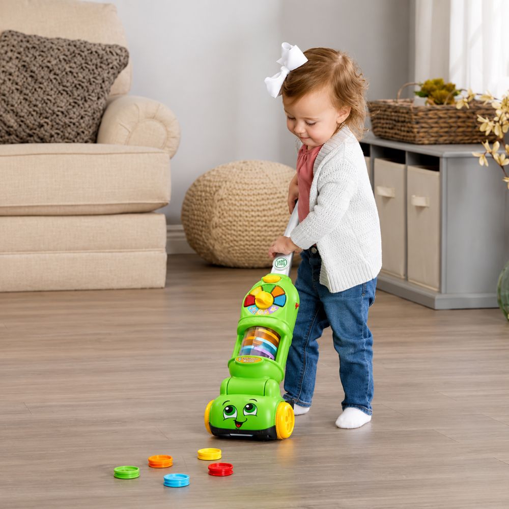A toddler girl pushes a bright green toy walker with colorful buttons and wheels in a cozy living room setting.
