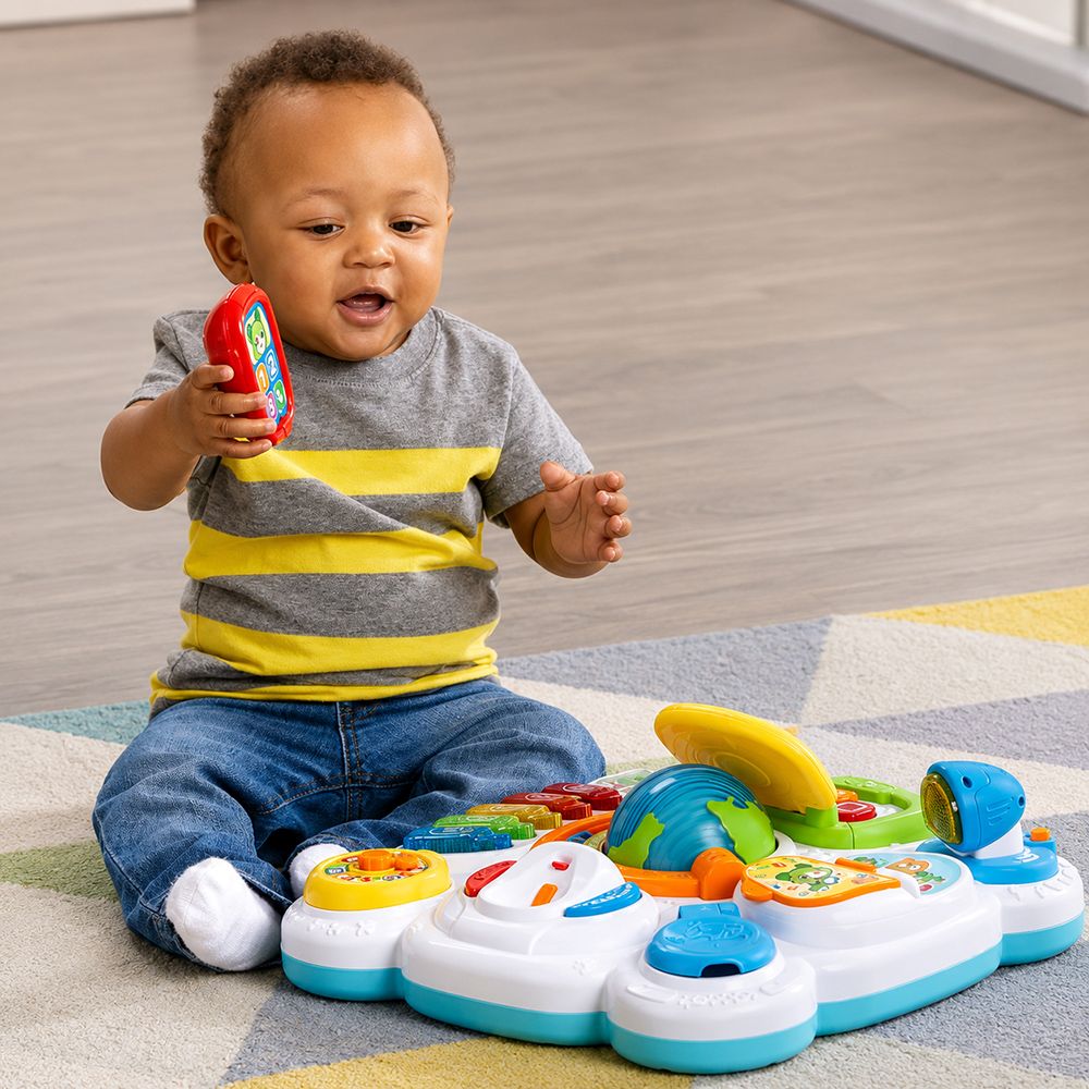 A baby sitting on a colorful rug plays with a white, blue, and yellow sensory activity mat on a wood floor. The baby is wearing a gray and yellow striped t-shirt and blue jeans, holding a red toy in one hand.