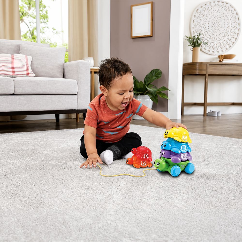 A toddler sits on a light gray rug in a bright living room, playing with a colorful stacking toy and pulling a red toy phone with a yellow cord.