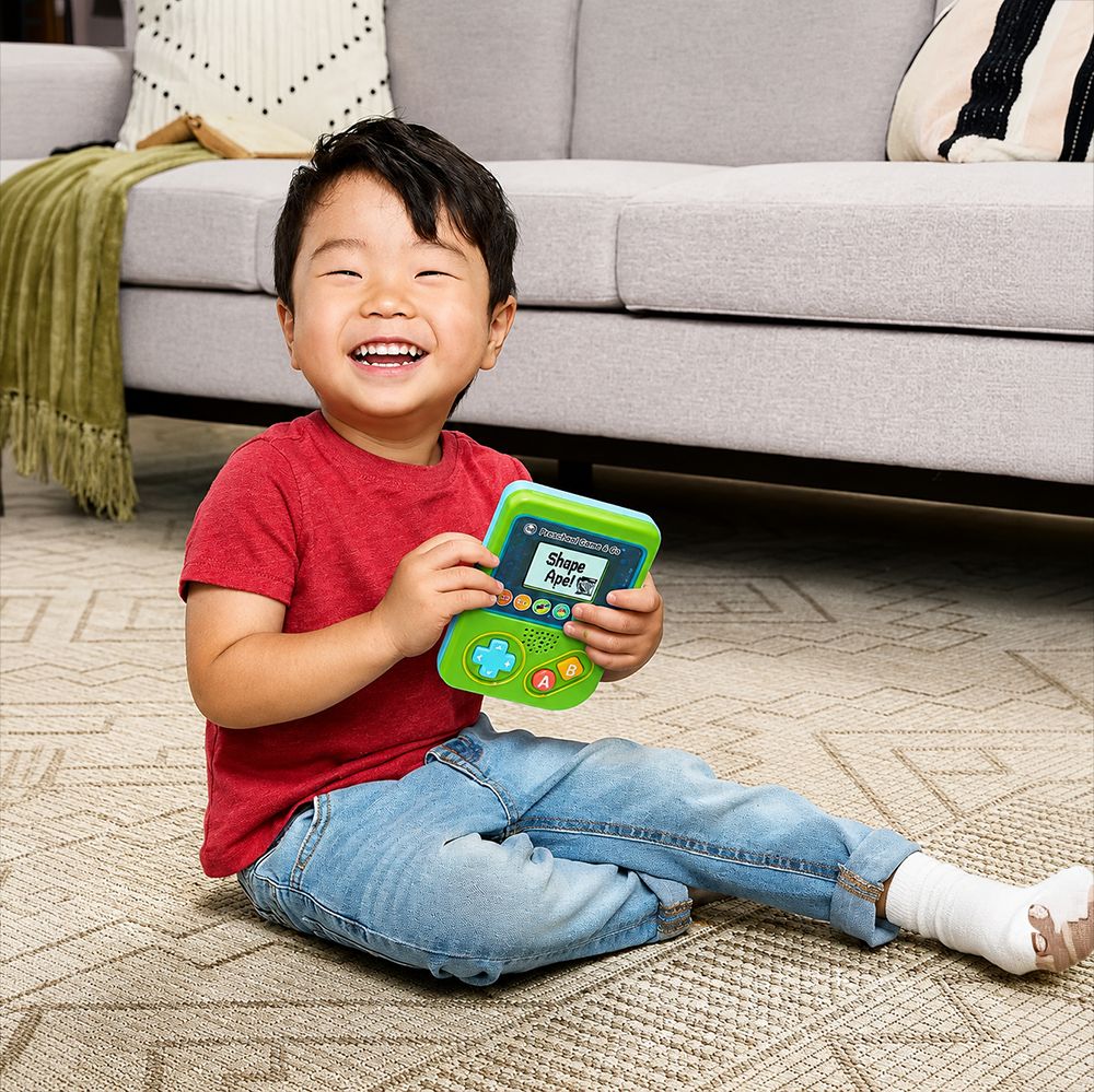 A young boy sitting on a patterned carpet in front of a gray sofa, smiling while holding a green and blue handheld toy game console.