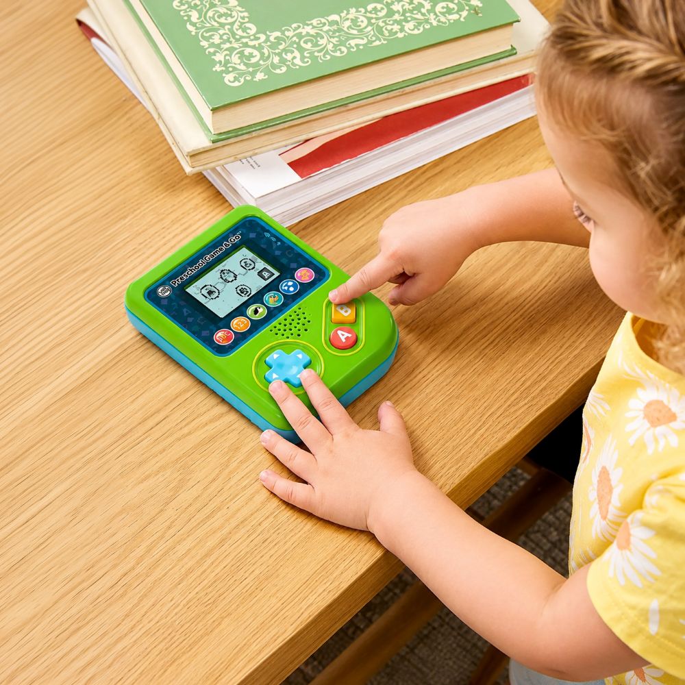 A young child in a yellow sunflower shirt interacts with a colorful green and blue touch learning toy on a wooden table, with books stacked in the background.